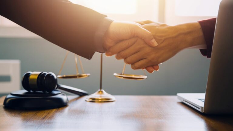 Lawyers shaking hands across a desk with a gavel and scales of justice, symbolizing a successful legal agreement.
