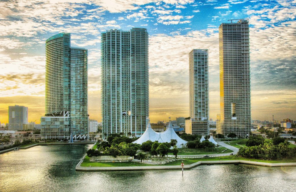 Panoramic view of modern high-rise buildings next to a waterfront with a white pavilion, at sunset, in a cityscape resembling Miami.”